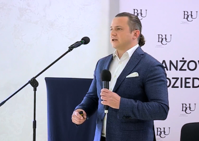 Prelegent Jakub Wróblewski Ekspert ds Badań i Analiz SRK OZiS przedstawia raport z badań przy stojącym mikrofonie na tle ścianki z logo Branżowego Centrum Umiejętności