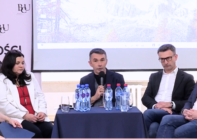 Uczestnicy panelu konferencyjnego siedzą przy stole i biorą udział w dyskusji, w tle ekran projekcyjny i branding BCU