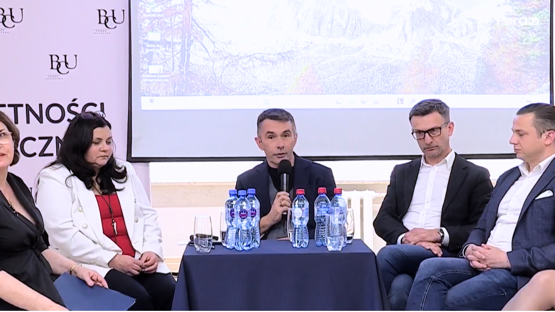 Uczestnicy panelu konferencyjnego siedzą przy stole i biorą udział w dyskusji, w tle ekran projekcyjny i branding BCU