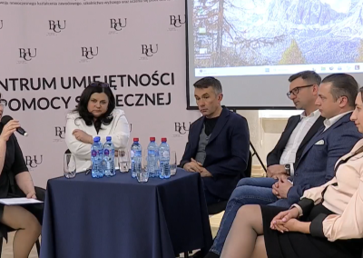 Panel dyskusyjny podczas konferencji w Branżowym Centrum Umiejętności w dziedzinie pomocy społecznej; prowadząca z mikrofonem rozmawia z uczestnikami siedzącymi przy stole.