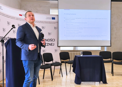 Prelegent prezentuje wyniki badania przy ekranie projekcyjnym podczas konferencji w Branżowym Centrum Umiejętności; obok stoją puste krzesła panelowe i stolik