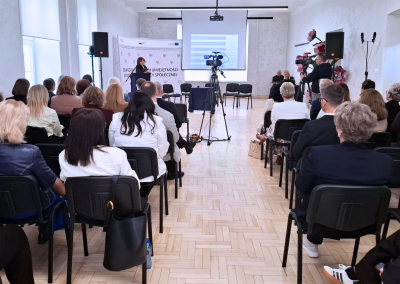 Sala konferencyjna podczas ogólnopolskiej konferencji o pomocy społecznej, widok od tyłu na licznie zgromadzoną publiczność, scenę z ekranem projekcyjnym oraz kamerę rejestrującą wydarzenie.