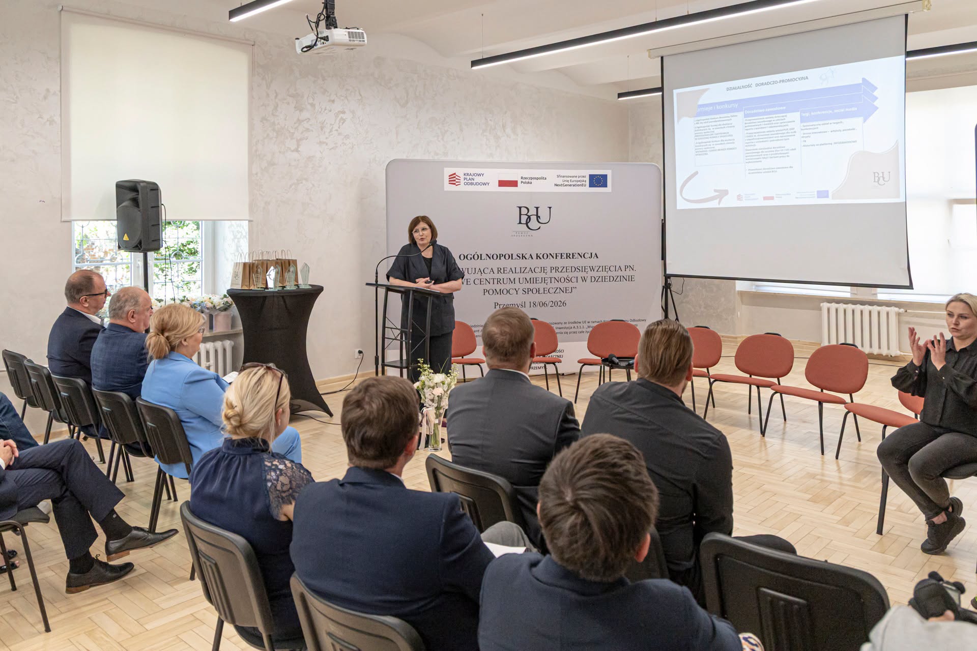 Uczestnicy panelu konferencyjnego siedzą przy stole i biorą udział w dyskusji, w tle ekran projekcyjny i branding BCU